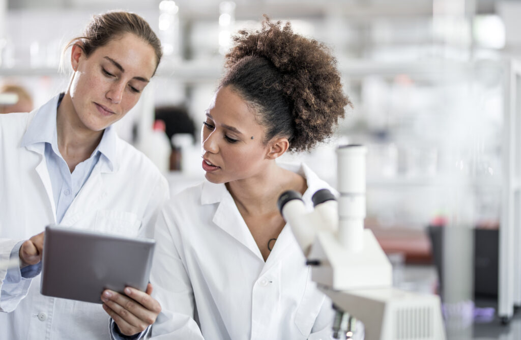 Zwei Frauen arbeiten in einem Labor, eine hält ein Tablet, die andere schaut auf den Bildschirm.