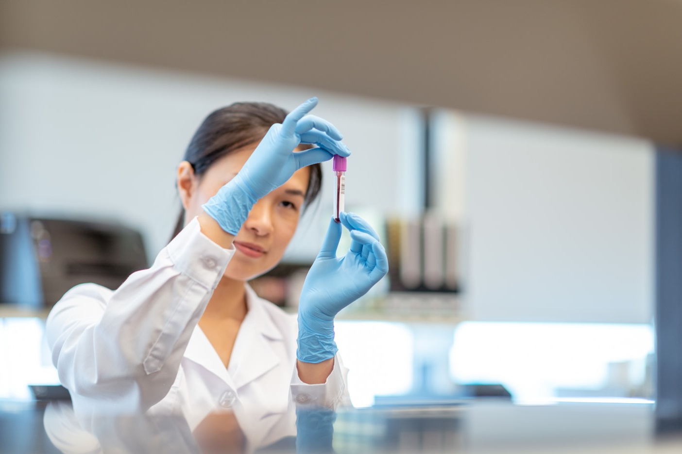 Une chimiste travaillant seule dans un laboratoire. La femme tient un petit flacon de sang et analyse l’échantillon médical. Elle porte une blouse de laboratoire et des gants de protection.