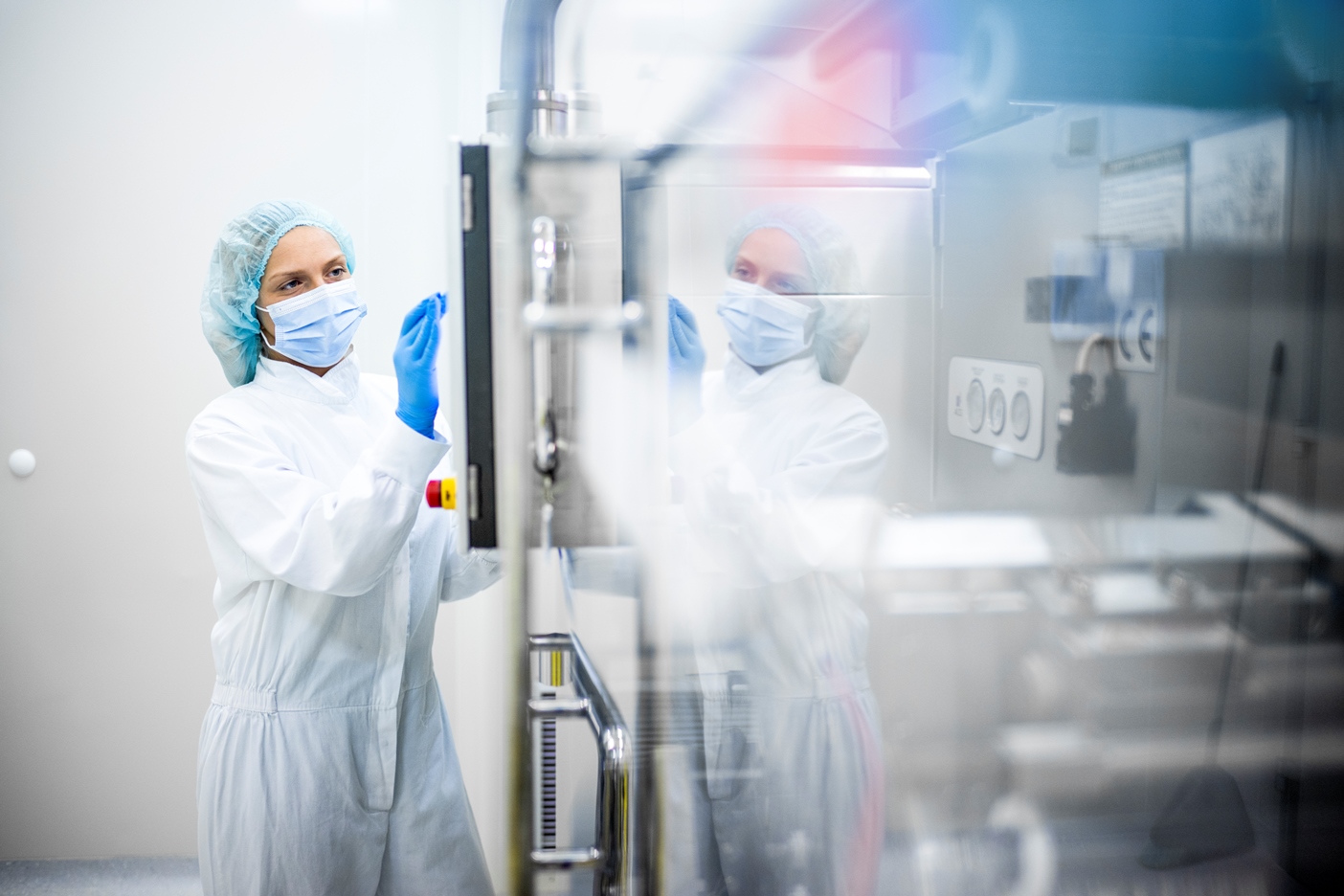 Image d’une femme portant des vêtements de sécurité appropriés en train de travailler sur l’écran d’une grande machine complexe dans l’industrie pharmaceutique, son reflet étant visible sur la machine.